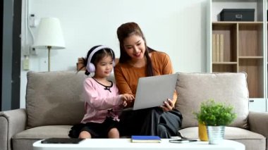 Neşeli anne ve kız kanepede güzel vakit geçiriyorlar, gülümsüyorlar ve kulaklık ve dizüstü bilgisayar kullanıyorlar. Aile teması için mükemmel.