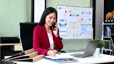 Dizüstü bilgisayarda çalışan ve ofisteki akıllı telefondan konuşan Asyalı genç iş kadını.