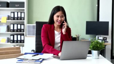 Dizüstü bilgisayarda çalışan ve ofisteki akıllı telefondan konuşan Asyalı genç iş kadını.
