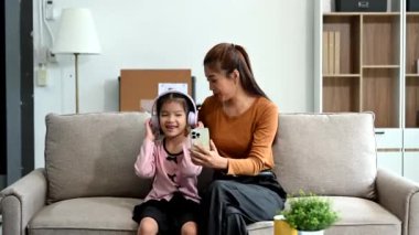 Neşeli anne ve kız kanepede güzel vakit geçiriyorlar, gülümsüyorlar ve kulaklık ve akıllı telefon kullanıyorlar. Aile teması için mükemmel.