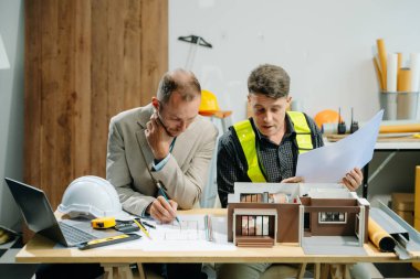 Çalışma masasında mimari tartışan, dizüstü bilgisayar kullanan ve planlara bakan meslektaşlar.