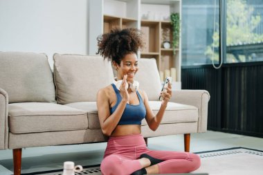 Atletizm sporu yapan aktif bir kadın evde yoga minderinde oturup, akıllı telefondan video görüşmesi yapıyor. Modern ev ortamında sağlıklı ve aktif bir yaşam tarzının göstergesi..