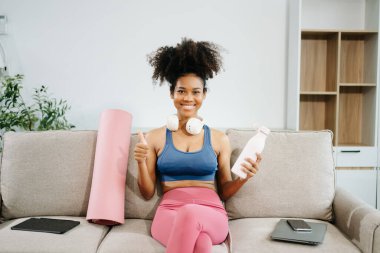 Spor salonundaki aktif mutlu kadın yoga yaptıktan sonra koltukta oturup başparmağını kaldırarak su şişesini tutuyor. Modern ev ortamında sağlıklı ve aktif bir yaşam tarzının göstergesi..