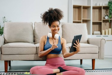 Spor salonundaki aktif kadın evde yoga minderinde oturup tablet kullanıyor. Modern ev ortamında sağlıklı ve aktif bir yaşam tarzının göstergesi..