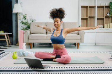 Afrika kökenli Amerikalı spor kadın yoga yapıyor ve dizüstü bilgisayarında eğitim görürken esniyor evdeki oturma odasında eğitim görüyor.