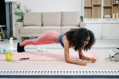 Modern bir ev ortamında yoga minderinde tahta pozisyonu alan genç bir kadın. Sağlığını, motivasyonunu ve sağlıklı aktif bir yaşam tarzını gösteriyor. 