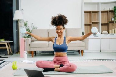 Modern bir ev ortamında çevrimiçi öğretim görevlileri izleyen ve halterlerle egzersiz yapan genç bir kadın. Sağlığını, motivasyonunu ve sağlıklı aktif bir yaşam tarzını gösteriyor. 