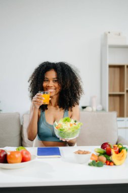 Modern bir mutfakta gülümseyen Afrikalı Amerikalı kadın taze meyve, sebze, blender ve portakal suyu ile temiz yeme, sağIık ve sağlıklı bir yaşam tarzını teşvik ediyor. 