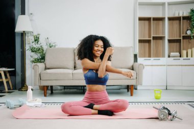 Genç, Afro-Amerikan bir kadın modern bir ev ortamında yoga minderinin üzerinde esneme egzersizi yapıyor. Sağlığını, motivasyonunu ve sağlıklı aktif bir yaşam tarzını gösteriyor. 