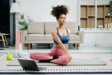 İnternette yoga yapmak için dizüstü bilgisayar kullanan ve modern ev ortamında paspas ve spor malzemeleriyle esneme hareketleri yapan genç bir kadın. Sağlığını, motivasyonunu ve sağlıklı aktif bir yaşam tarzını gösteriyor. 