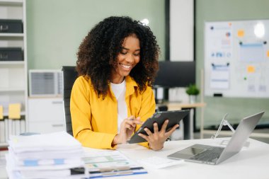 Gülümseyen Afrikalı Amerikalı iş kadını modern ofiste tablet ve dizüstü bilgisayarla çalışıyor. 