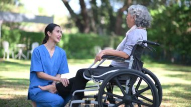 Genç hemşire, tekerlekli sandalyedeki yaşlı Asyalı kadınla ilgileniyor, hastane bahçesinde bacak egzersizi yapıyor.