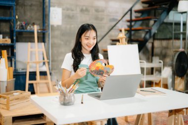 Neşeli bir Asyalı kadın sanatçı canlı bir stüdyoda dizüstü bilgisayar ve palet kullanıyor. Yaratıcılık ve sanatsal ilham temaları için mükemmel