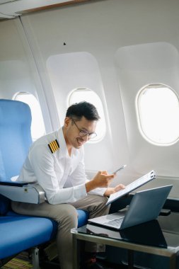 Dizüstü bilgisayarda çalışırken rahat koltukta oturan ve akıllı telefon kullanan Asyalı erkek pilot. Tarz içinde seyahat edin, zarafetle çalışın.