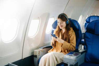 Asyalı bir kadın birinci sınıf uçak koltuğunda kulaklık takıyor ve akıllı telefon kullanıyor. Lüks, konfor ve zarafet sergiliyor. Havacılık, turizm ve iş hayatı için mükemmel. 