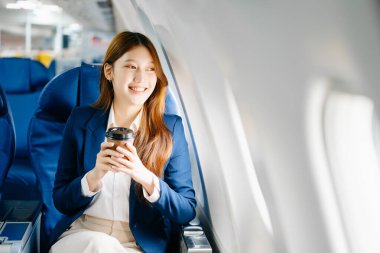 Birinci sınıf uçak koltuğunda oturan ve kahve içen Asyalı bir kadın lüks, rahat ve şık bir seyahat sergiliyor. Havacılık, turizm ve iş hayatı için mükemmel. 