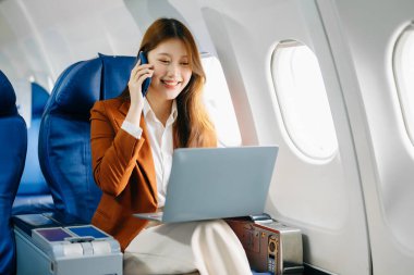 Asyalı bir kadın birinci sınıf uçak koltuğunda ve dizüstü bilgisayar kullanıyor ve akıllı telefonla konuşuyor, lüks, rahat ve zarif bir seyahat sergiliyor. Havacılık, turizm ve iş hayatı için mükemmel. 