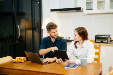 Modern bir çift, rahat bir mutfak ortamında dizüstü bilgisayar, defter ve kahvaltıyla birlikte kahve içerken iş birliği yapıyor. Yaşam tarzı, takım çalışması ve günlük verimliliğin mükemmel bir karışımı.