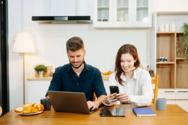 Modern bir çift, rahat bir mutfak ortamında dizüstü bilgisayar, defter ve kahvaltıyla birlikte kahve içerken iş birliği yapıyor. Yaşam tarzı, takım çalışması ve günlük verimliliğin mükemmel bir karışımı.