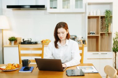 Evde otururken dizüstü bilgisayar kullanan bir kadın. Genç bir kadın mutfakta oturuyor ve dizüstü bilgisayarla çalışıyor..