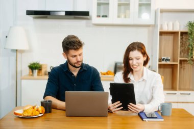 Modern bir çift, rahat bir mutfak ortamında dizüstü bilgisayar, defter ve kahvaltıyla birlikte kahve içerken iş birliği yapıyor. Yaşam tarzı, takım çalışması ve günlük verimliliğin mükemmel bir karışımı.