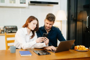 Modern bir çift, rahat bir mutfak ortamında dizüstü bilgisayar, defter ve kahvaltıyla birlikte kahve içerken iş birliği yapıyor. Yaşam tarzı, takım çalışması ve günlük verimliliğin mükemmel bir karışımı.