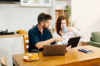 Modern bir çift, rahat bir mutfak ortamında dizüstü bilgisayar, defter ve kahvaltıyla birlikte kahve içerken iş birliği yapıyor. Yaşam tarzı, takım çalışması ve günlük verimliliğin mükemmel bir karışımı.