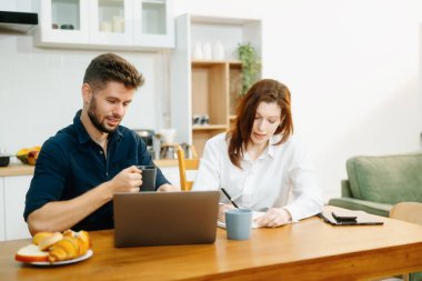 Modern bir çift, rahat bir mutfak ortamında dizüstü bilgisayar, defter ve kahvaltıyla birlikte kahve içerken iş birliği yapıyor. Yaşam tarzı, takım çalışması ve günlük verimliliğin mükemmel bir karışımı.