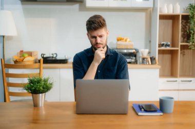 İş adamı evde otururken dizüstü bilgisayar kullanıyor. Genç adam mutfakta oturuyor ve dizüstü bilgisayarla çalışıyor..