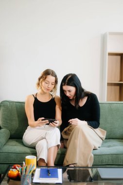 İki neşeli kadın yeşil bir koltukta oturmuş, akıllı telefonları kullanarak şık bir ev ortamında fikir alışverişinde bulunuyorlar. Canlı takım çalışmasını, yaratıcılığı ve genç yetişkinlerle öğrenmeyi keşfedin..