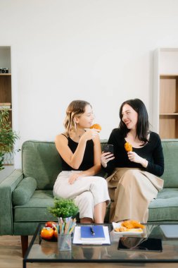 İki neşeli kadın yeşil bir kanepede oturmuş tavuk yiyor ve şık bir evde akıllı telefon kullanıyor. Takım çalışması çalışma alanı temaları için mükemmel