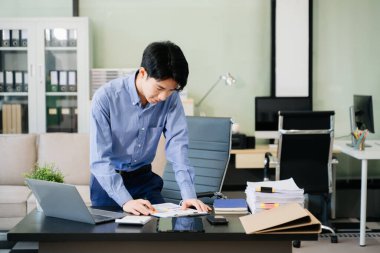 Genç iş adamı ofiste dizüstü bilgisayarla çalışıyor ve kağıda not alıyor..