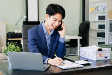 Genç iş adamı ofiste dizüstü bilgisayarla, tabletle çalışıyor ve kağıda not alıyor..