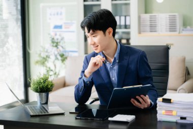 Genç iş adamı ofiste dizüstü bilgisayarla, tabletle çalışıyor ve kağıda not alıyor..