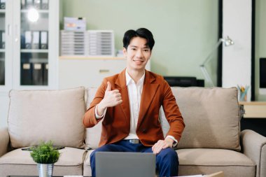 Dizüstü bilgisayarı olan Asyalı girişimci şık bir ofiste koltukta oturuyor ve başparmağını gösteriyor. İş için mükemmel, üretkenlik teması