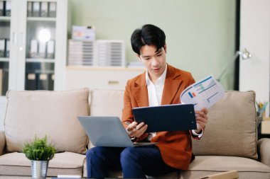 Asyalı girişimci işine odaklanmış, şık bir ofiste koltukta dizüstü bilgisayarla belgeleri analiz ediyor. İş için mükemmel, verimlilik için.