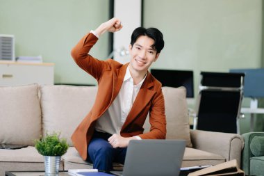 Kolunu kaldırmış Asyalı girişimci şık bir ofiste kanepede dizüstü bilgisayar kullanıyor. İş için mükemmel, üretkenlik teması