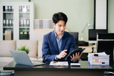 Genç iş adamı ofiste dizüstü bilgisayar ve tabletle çalışıyor.