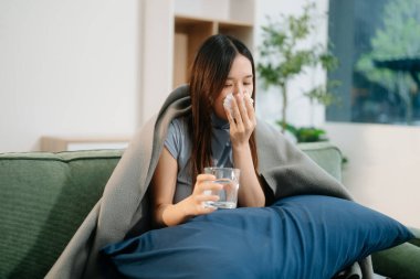 Genç Asyalı kadın grip belirtilerinden muzdarip, ellerinde hapşıran ve bir bardak su tutan, kanepenin üzerindeki battaniyeyle kaplanmış. Hastalık, sağlık ve iyileşme kavramı. Evde kal, sağlığına dikkat et.