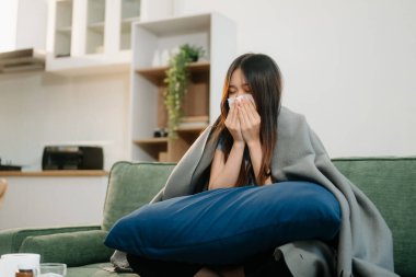 Genç Asyalı kadın grip belirtilerinden muzdarip, hapşırıyor ve kanepede battaniyeyle kaplanıyor. Hastalık, sağlık ve iyileşme kavramı. Evde kal, sağlığına dikkat et.