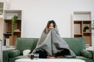 Kanepede battaniyeyle kaplanmış, grip belirtileri gösteren genç bir Asyalı kadın. Hastalık, sağlık ve iyileşme kavramı. Evde kal, sağlığına dikkat et.