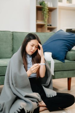 Grip belirtileri gösteren genç bir Asyalı kadın, elinde termometreyle kanepede battaniye tutuyor. Hastalık, sağlık ve iyileşme kavramı. Evde kal, sağlığına dikkat et.