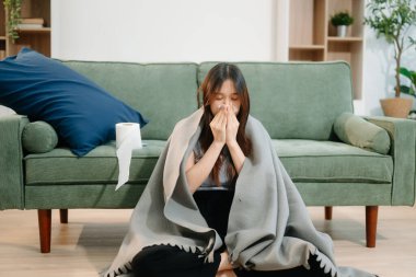 Genç Asyalı kadın grip belirtilerinden muzdarip, hapşırıyor ve kanepede battaniyeyle örtülmüş. Hastalık, sağlık ve iyileşme kavramı. Evde kal, sağlığına dikkat et.