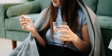 Genç Asyalı kadın grip belirtilerinden muzdarip, elinde termometre ve bir bardak su tutan, kanepede battaniyeyle yatan. Hastalık, sağlık ve iyileşme kavramı. Evde kal, sağlığına dikkat et.