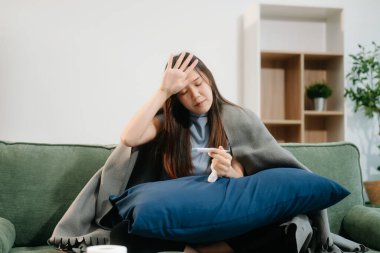 Grip belirtilerinden muzdarip genç bir Asyalı kadın elinde termometreyle kanepenin üzerinde battaniye tutuyor. Hastalık, sağlık ve iyileşme kavramı. Evde kal, sağlığına dikkat et.