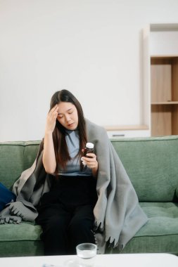 Grip belirtilerinden muzdarip genç bir Asyalı kadın elinde bir şişe hap, kanepenin üzerinde battaniyeyle. Hastalık, sağlık ve iyileşme kavramı. Evde kal, sağlığına dikkat et.