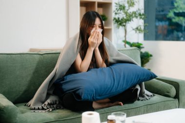 Genç Asyalı kadın grip belirtilerinden muzdarip, hapşırıyor, kanepenin üzerinde battaniyeyle örtülmüş. Hastalık, sağlık ve iyileşme kavramı. Evde kal, sağlığına dikkat et.