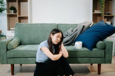 Depresyondaki Asyalı kadın oturma odasında kanepede oturmaktan endişe duyuyor. Hastalık, sağlık ve iyileşme kavramı.