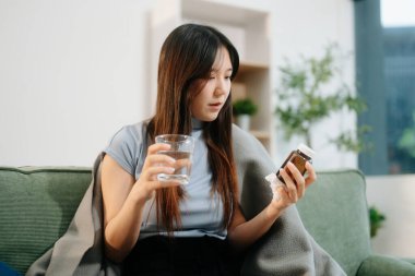 Genç Asyalı kadın grip belirtilerinden muzdarip, elinde bir şişe hap ve bir bardak su, kanepenin üzerinde battaniyeyle. Hastalık, sağlık ve iyileşme kavramı. Evde kal, sağlığına dikkat et.