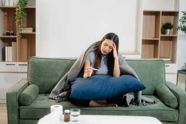 Grip belirtilerinden muzdarip genç bir Asyalı kadın elinde termometreyle kanepenin üzerinde battaniye tutuyor. Hastalık, sağlık ve iyileşme kavramı. Evde kal, sağlığına dikkat et.